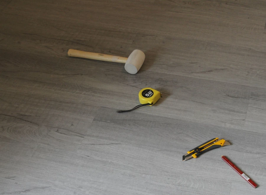 Gummihammer, Maßband, Cuttermesser und Bleistift auf einem grauen Vinylboden